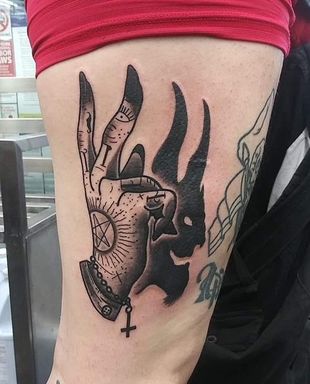 Hand with small, square tattoo of arrows and a heart. Gold ring on finger, black jacket, grey background.