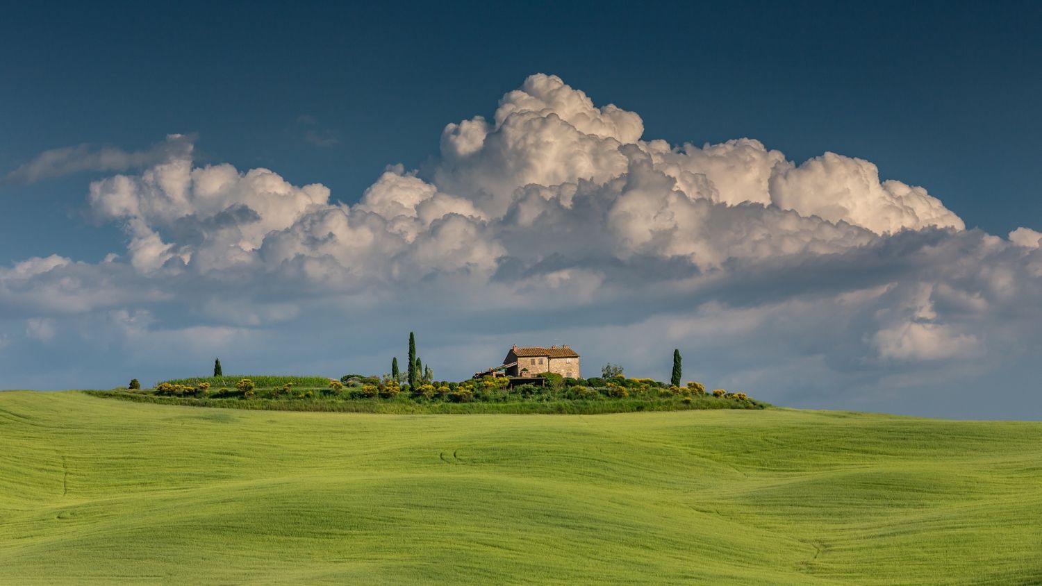 Rolling green Tuscan hills with a small building on a hilltop under fluffy white clouds.