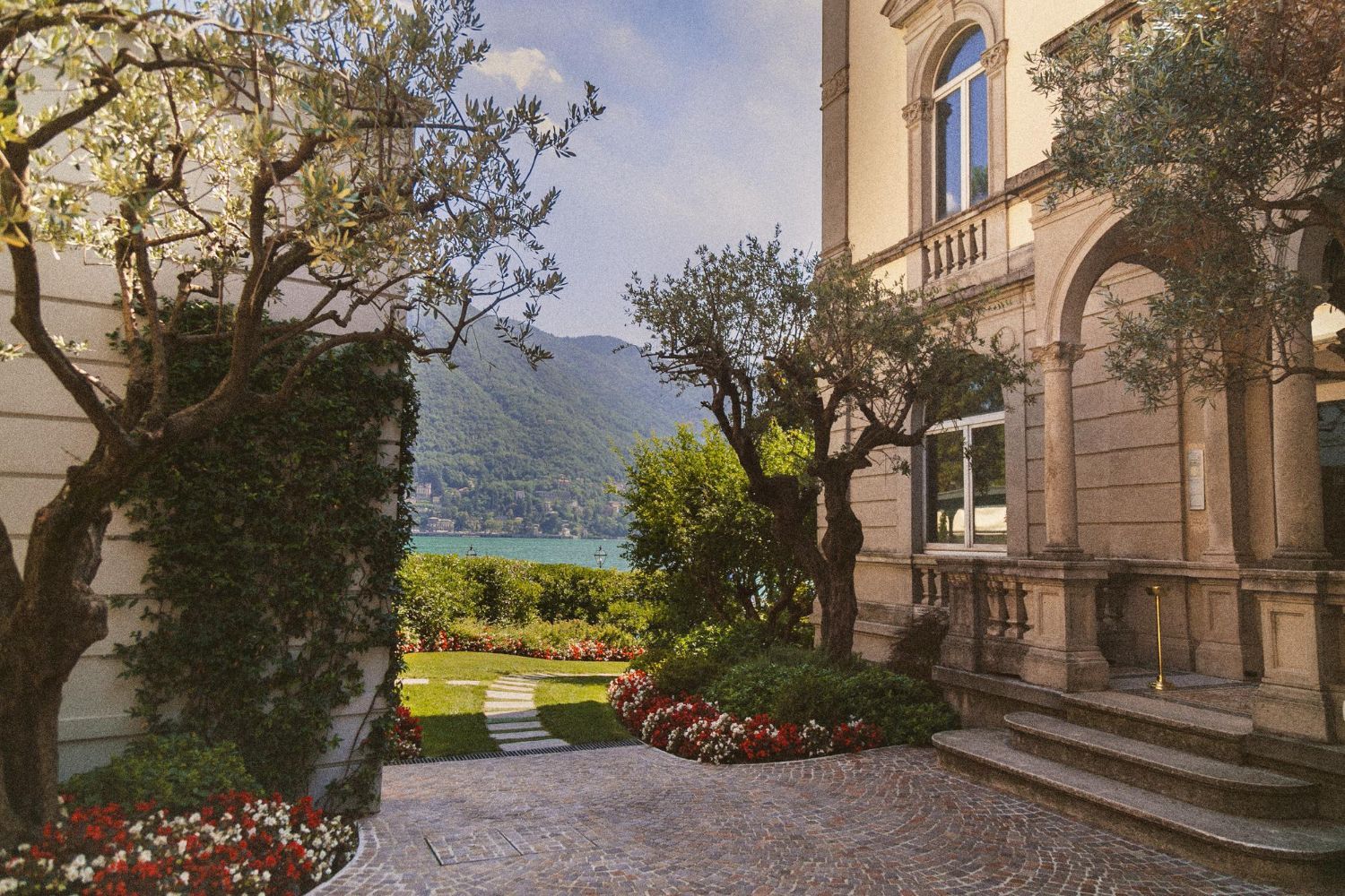 Elegant building exterior with arched entrance, stone patio, olive trees, and garden overlooking a lake.