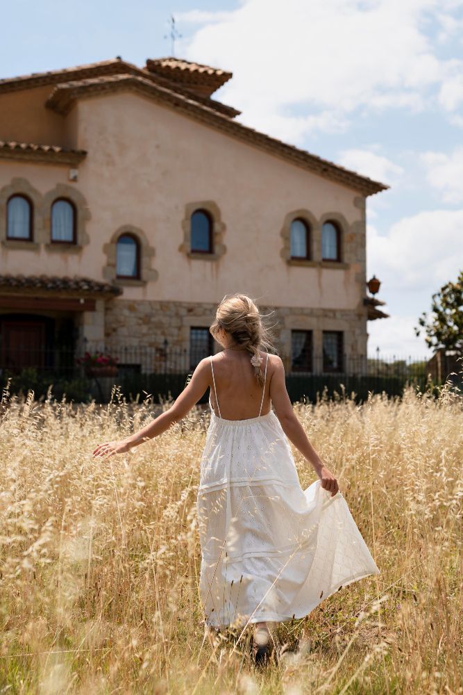Woman in white dress walks through tall dry grass toward a building with arched windows.