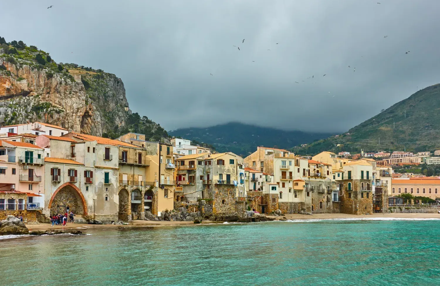 Coastal town of Cefalù, Sicily, with beige buildings, turquoise water, and a rocky mountain backdrop under a cloudy sky.