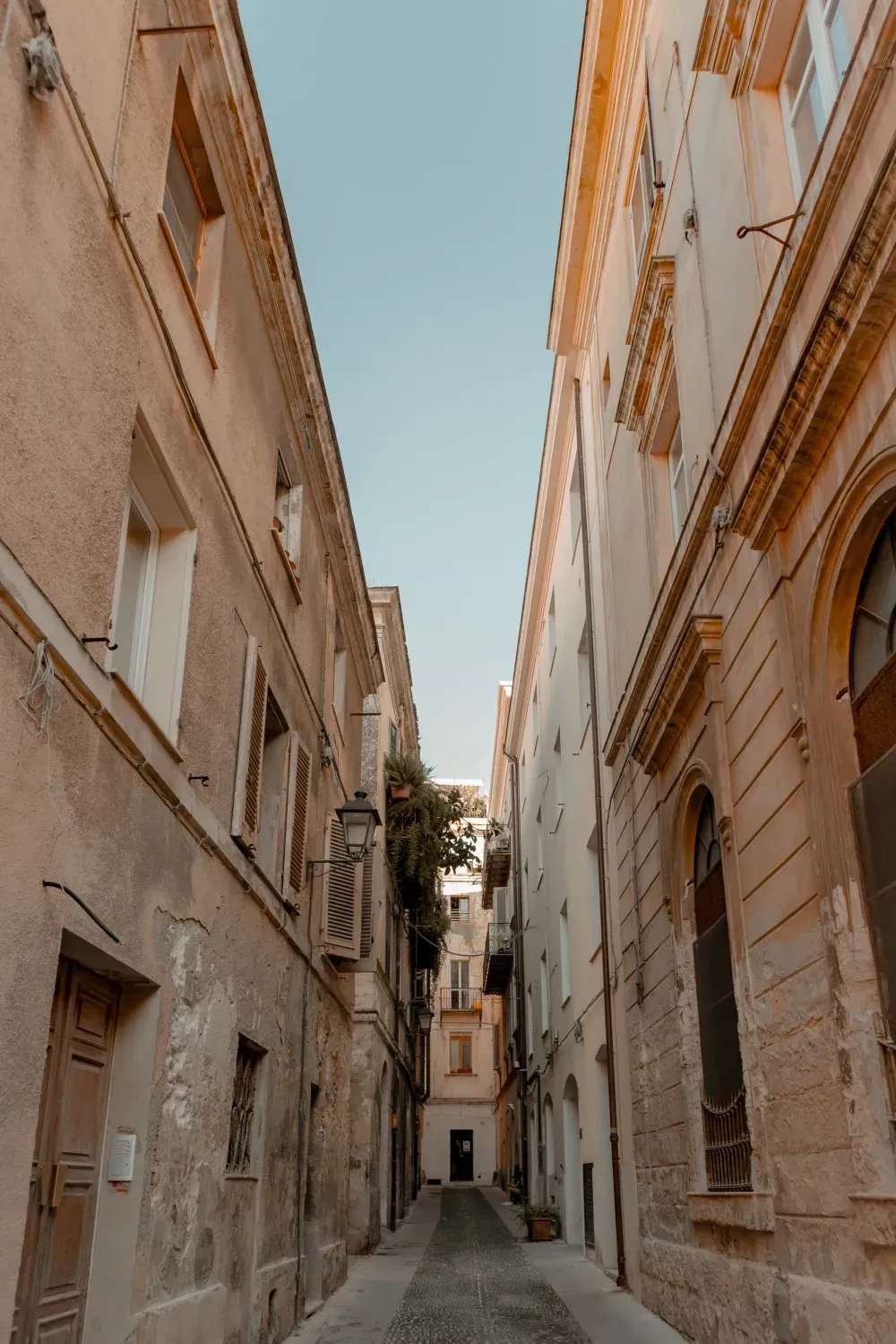 Narrow European cobblestone street between beige buildings, leading to a small doorway under a clear blue sky.