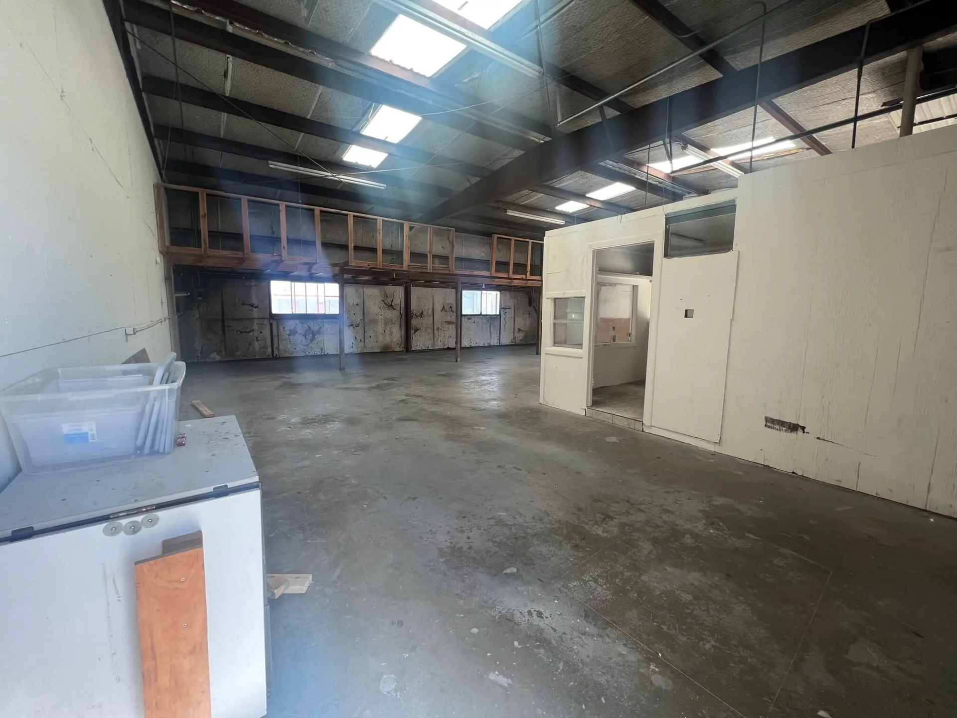 Empty industrial space with concrete floor, metal roof, and daylight from skylights. Storage or office area in the background.
