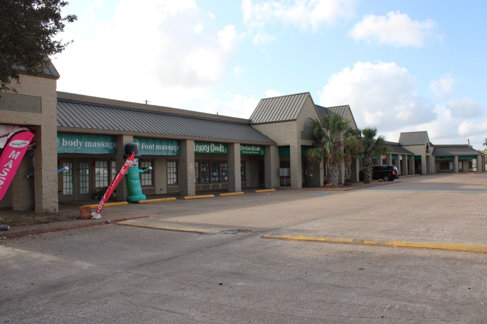 Shops in a strip mall with a parking lot. Light-colored brick, teal and green accents, and a blue sky.
