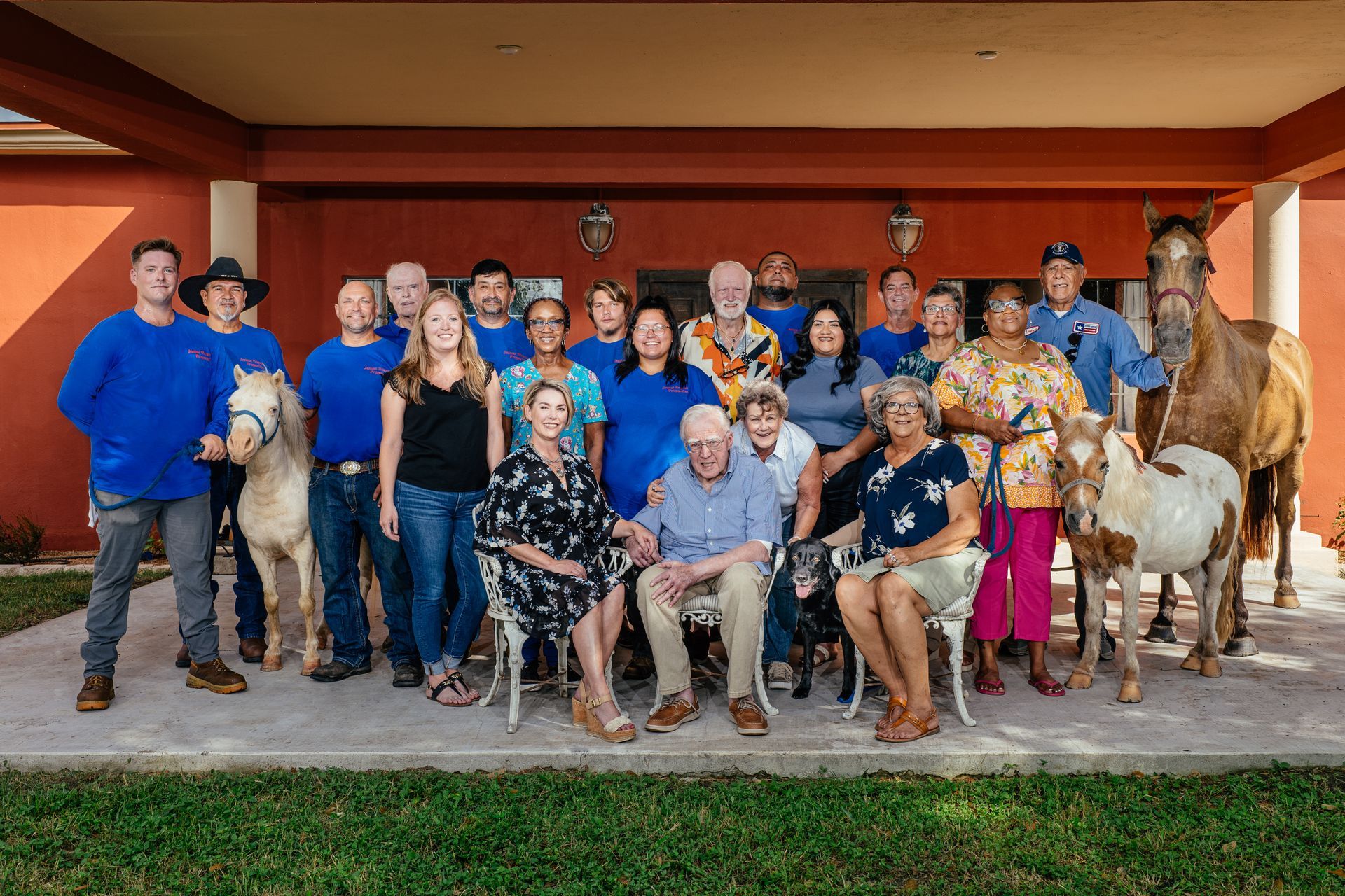 Group photo with people and horses on a porch. Many are wearing blue, with some sitting and others standing.