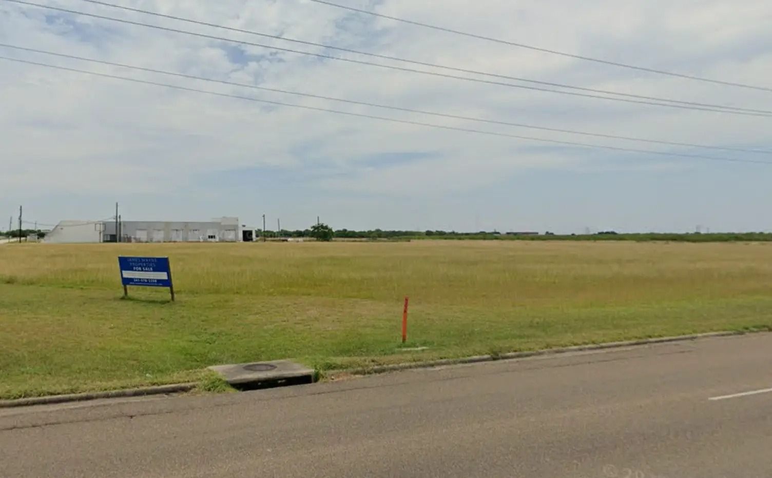 Grassy field with a building in the background under a cloudy sky. A sign and a sewer grate are in the foreground.