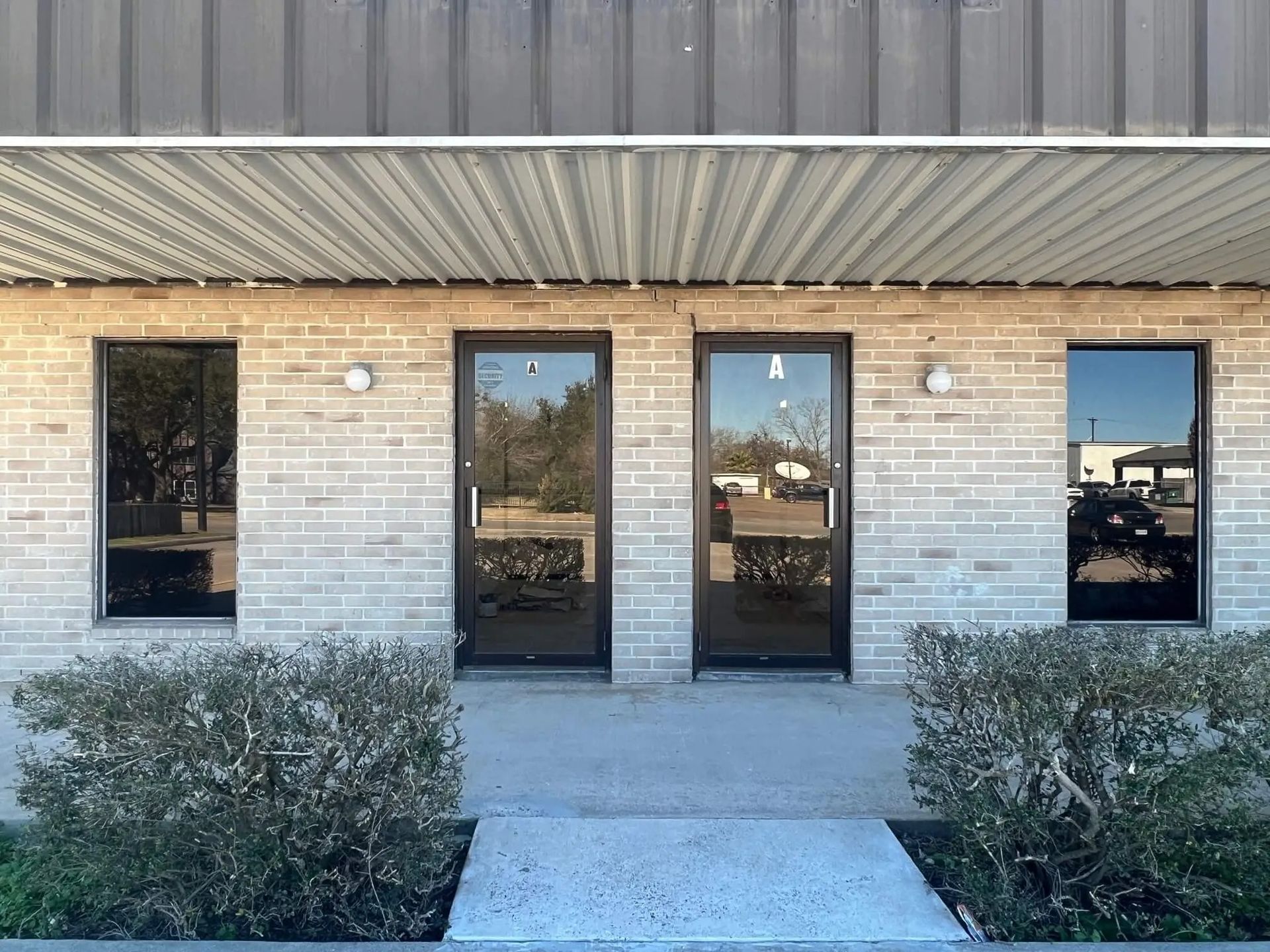 Exterior of a brick building with two glass doors and two windows, with bushes in front. Reflective glass shows surroundings.