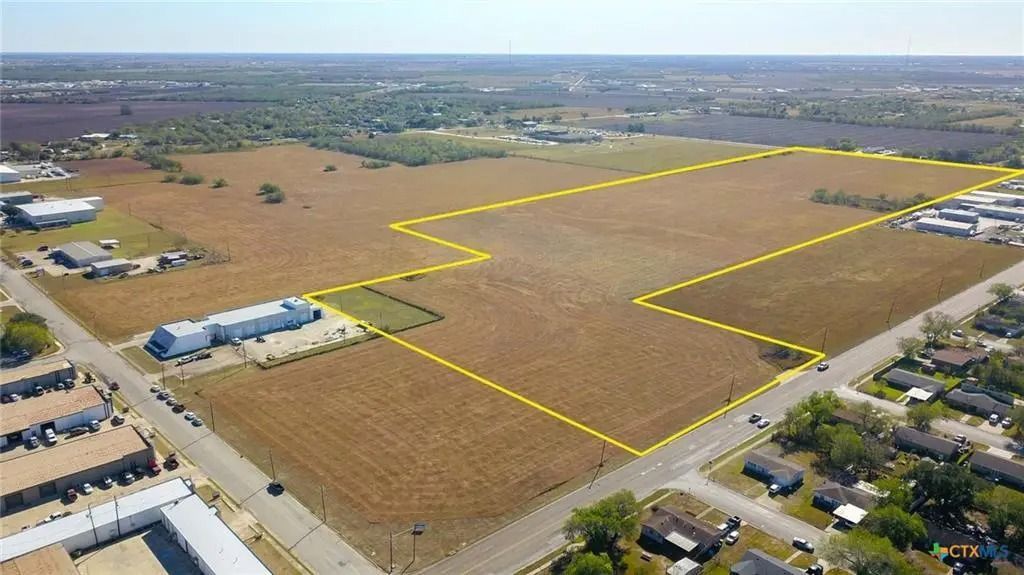 Aerial view of a large, vacant lot outlined in yellow, surrounded by roads, buildings, and houses under a bright sky.