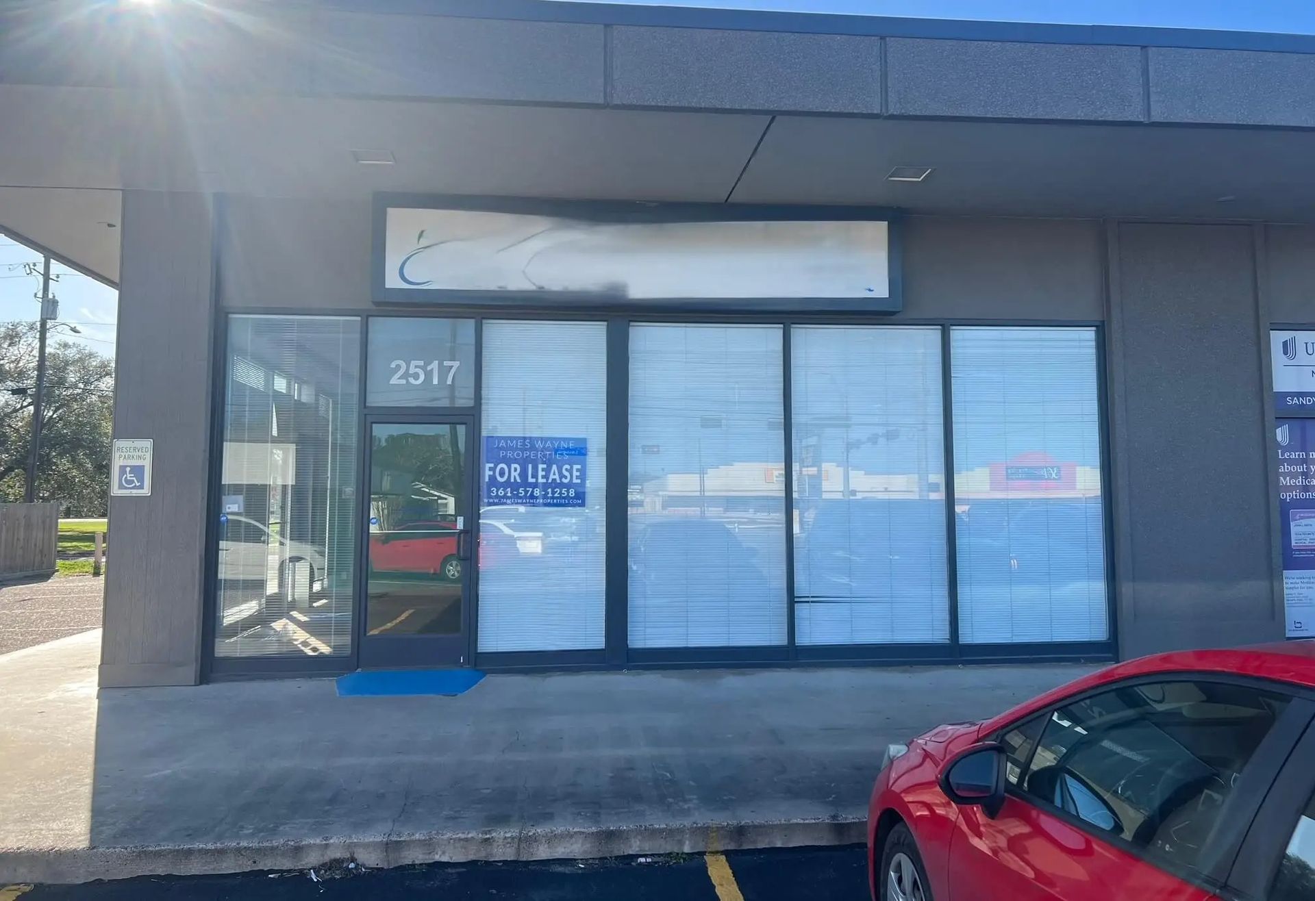Exterior of a commercial building with a "For Lease" sign in the window. The storefront is gray with a red car parked in front.