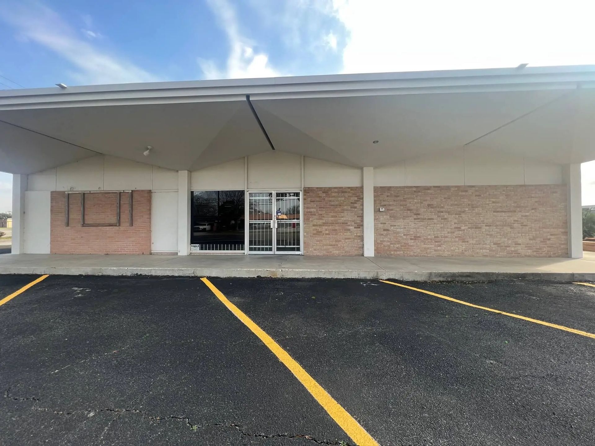 Exterior of a brick and concrete building with an overhanging roof and a security gate over the entrance, on an empty asphalt parking lot.