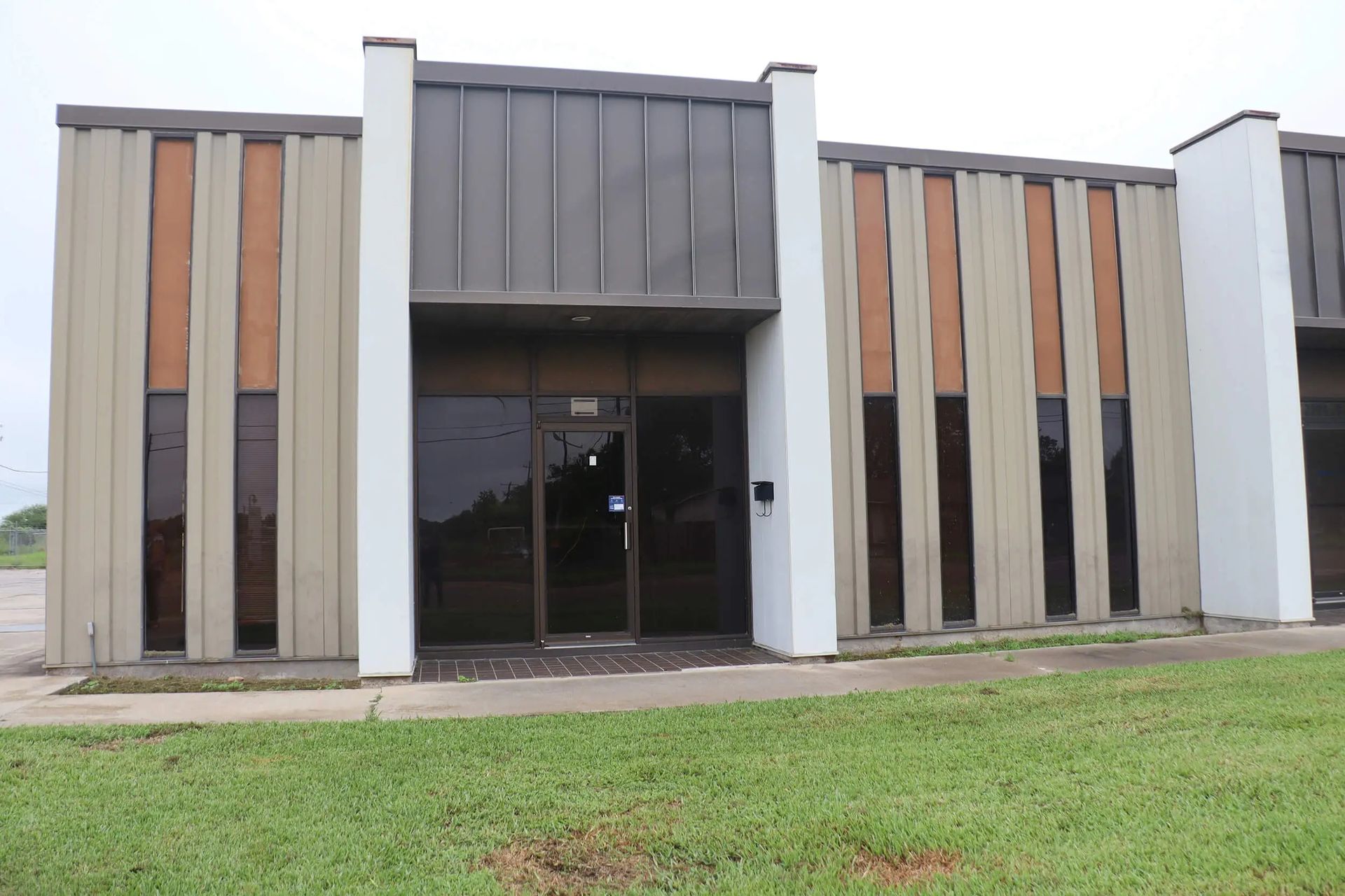 An abandoned building with vertical tan and orange panels, a dark roof, and glass doors. The building is bordered by white columns.
