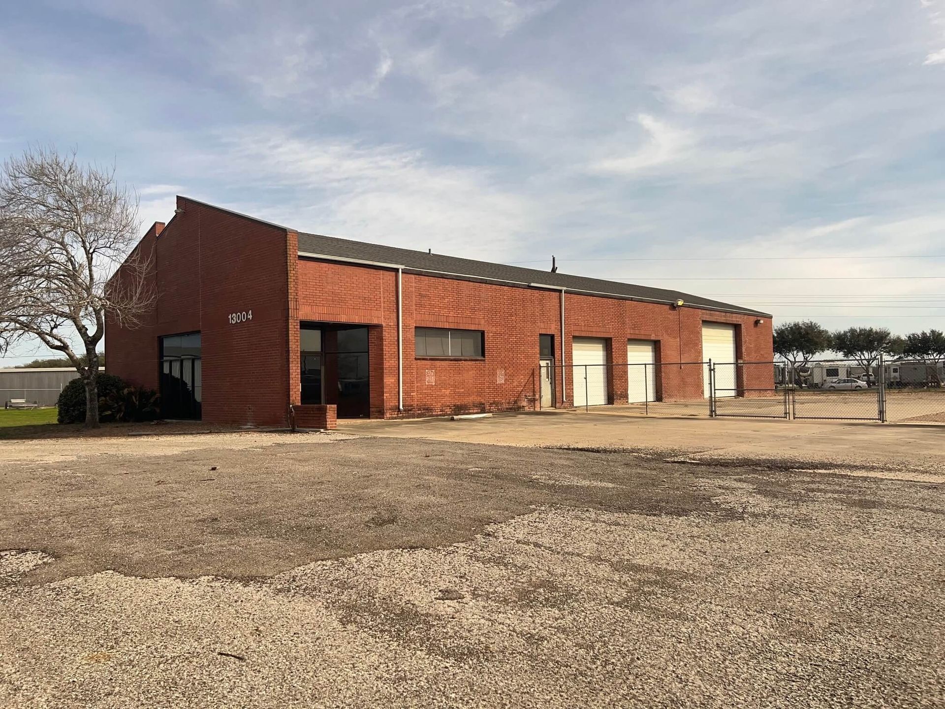 Brick building with three garage doors, a small office door, and a gravel parking area under a partly cloudy sky.