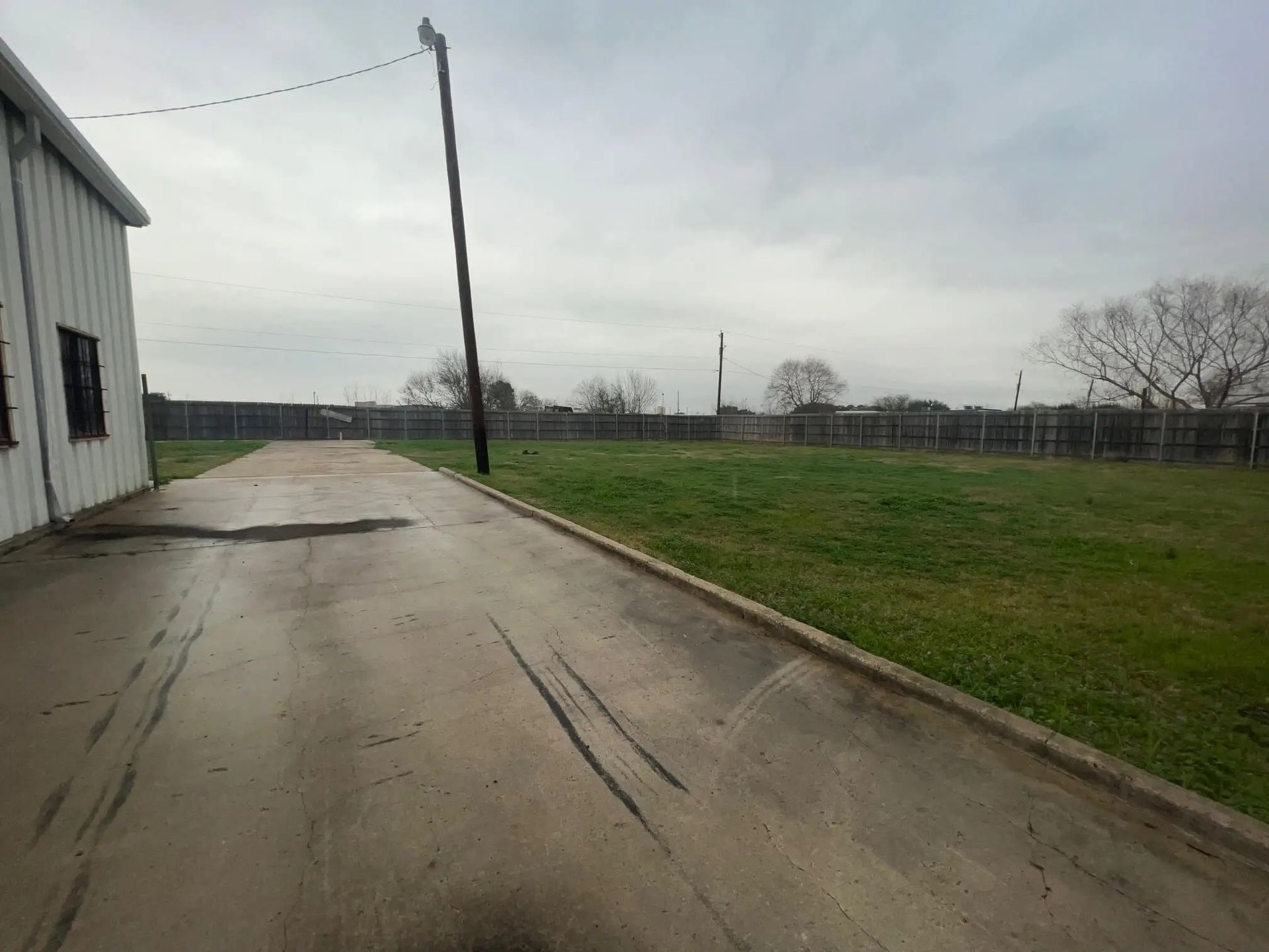 A gray, overcast day with a wet concrete driveway leading to a grassy field fenced in with a building on the left.