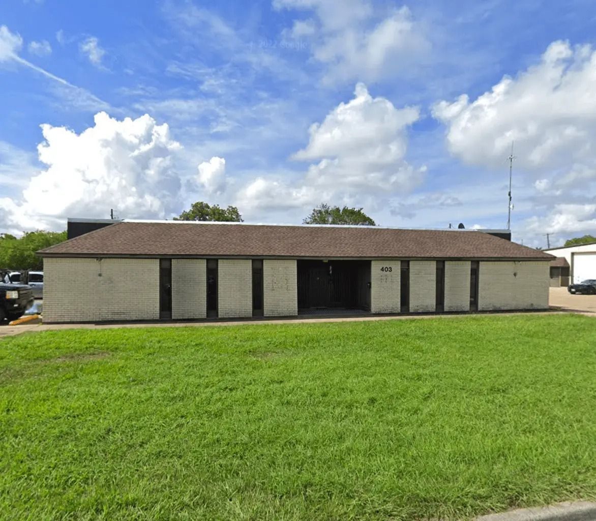A one-story, light-colored brick building with a brown roof sits behind a grassy lawn under a partly cloudy blue sky.