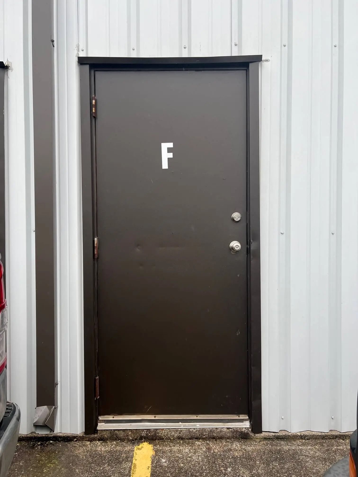 Brown door with the letter "F" on it, set in a white metal building. A yellow line and part of a car are visible.