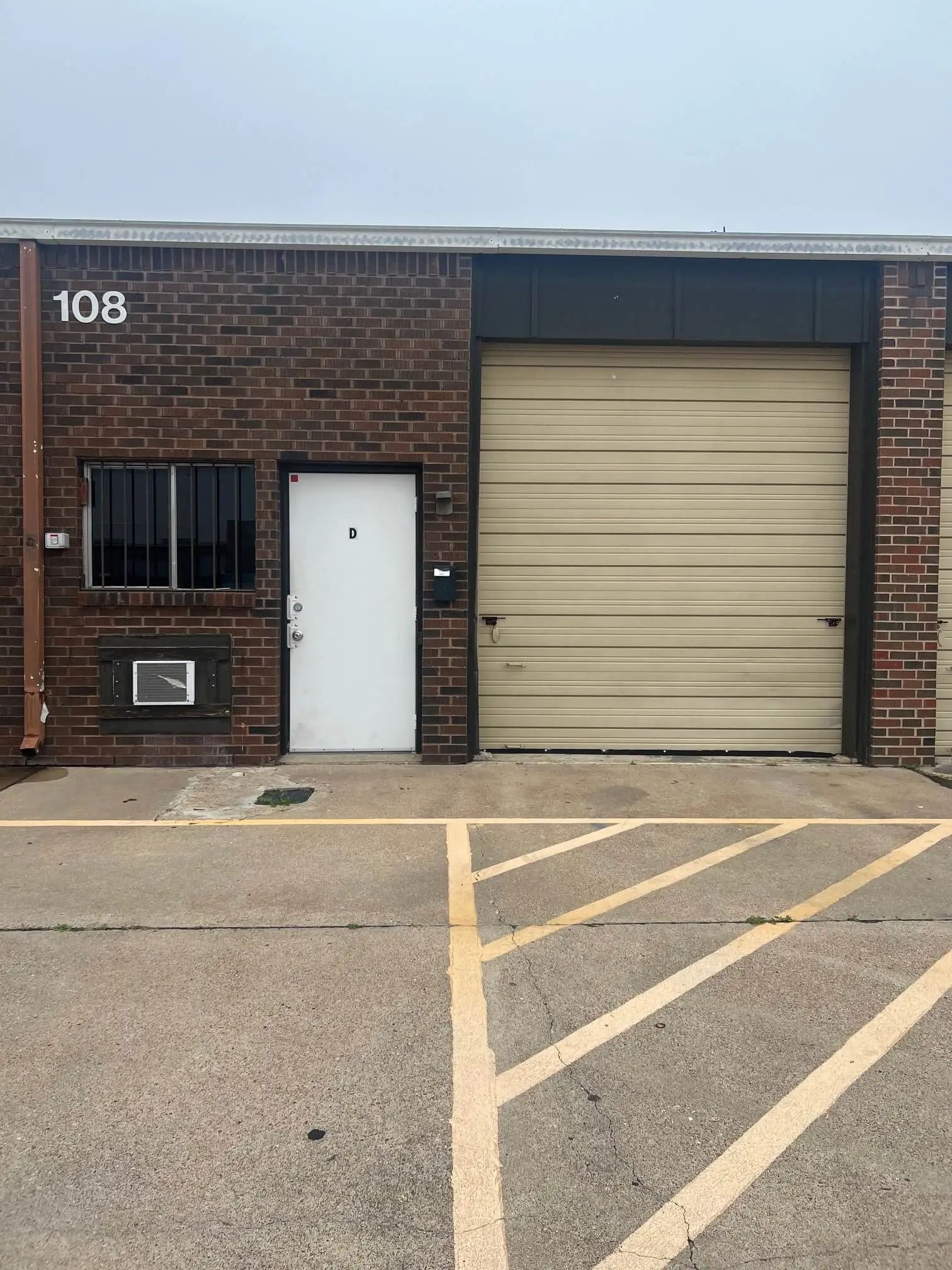 A brick building with unit number 108, a white door, a garage door, and a window. Yellow lines on the pavement in front.