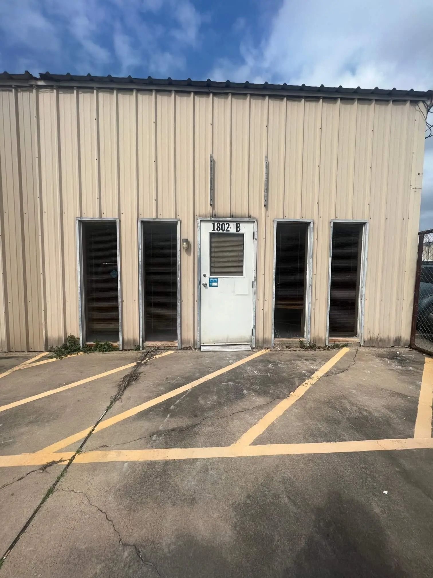 Exterior of a weathered, light-colored building with open doorways. Asphalt parking lot with yellow painted lines in front.
