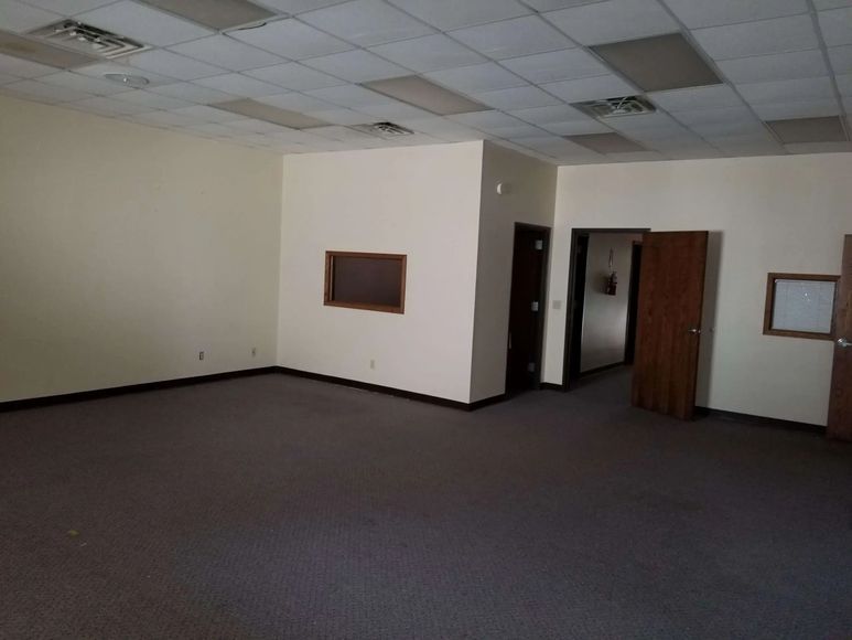 Empty office space with beige walls, brown carpet, and a drop ceiling. Two doorways lead to other rooms.