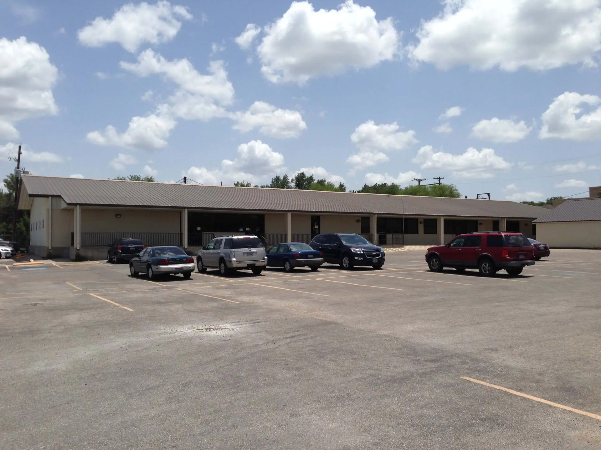 Cars parked in front of a one-story building with a tan facade on a sunny day with a cloudy sky.