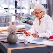 Senior woman with glasses works at her desk, using a phone and a pen.