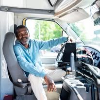Black man driving a vehicle, smiling. He wears a blue shirt in a bright setting.