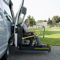 Man in wheelchair using a lift to enter a wheelchair-accessible van, outdoor setting.