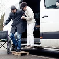 Man in wheelchair assisted by another person exiting a van using a wooden ramp.
