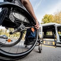 Person in wheelchair near a van with a ramp, on a paved road. Blue jeans, sunny day.