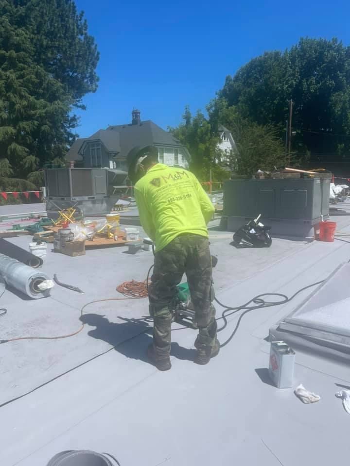 Worker in neon shirt repairing a white flat roof under a clear blue sky, with tools and equipment nearby.