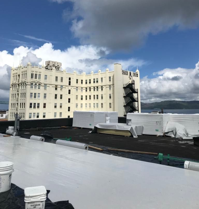 Rooftop with white roofing, stacked materials, and a cream building under a cloudy blue sky