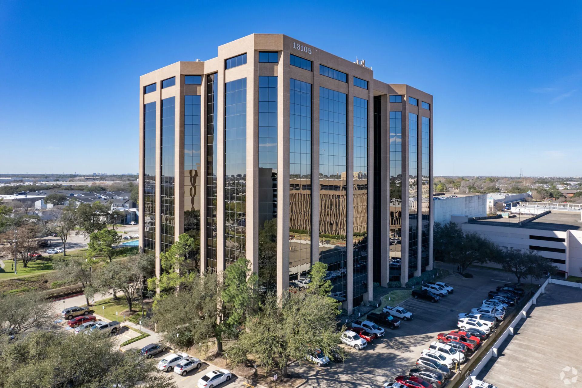 Tall building with mirrored blue windows and tan trim under a bright blue sky, framed by tree branches.