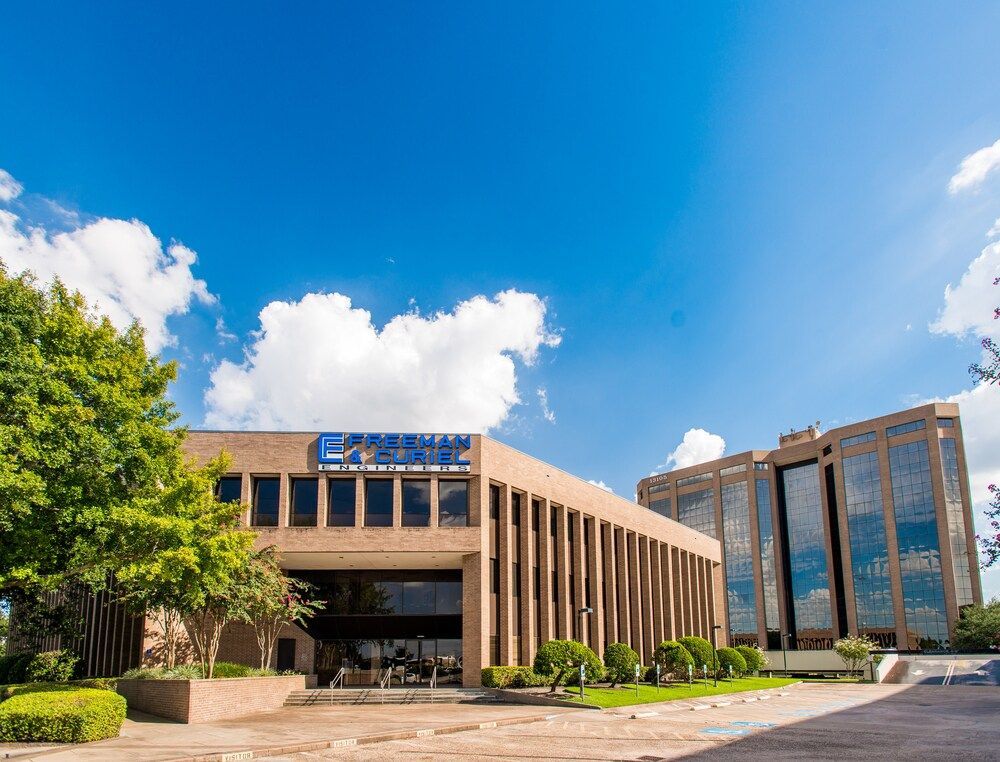 A large beige building with vertical window columns. Green bushes line the base; a blue sky with clouds in the background.