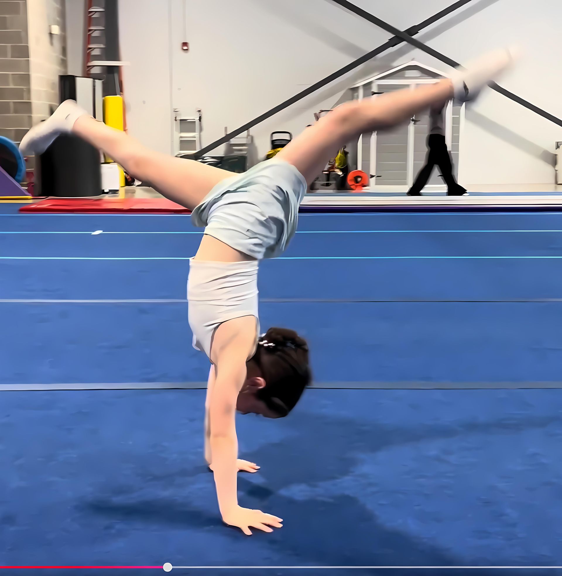 Young person doing a handstand on a blue mat, legs split in the air. Gym interior.