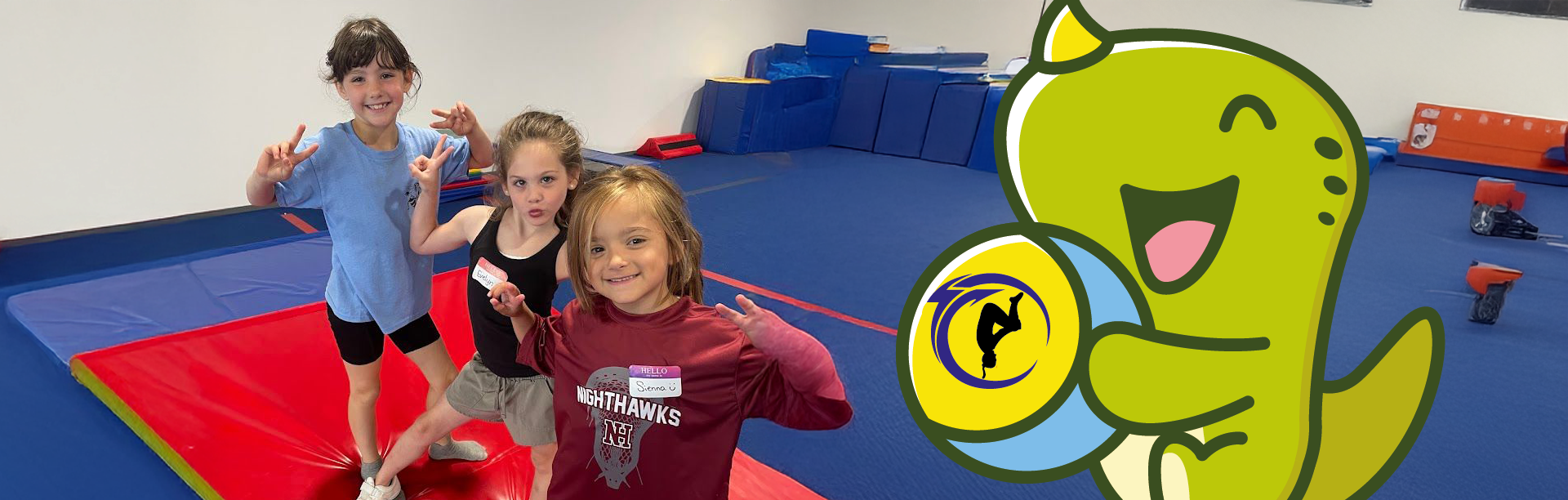 Three kids smiling and posing in a gym with a cartoon dinosaur holding a ball.