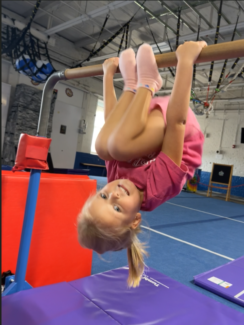 Child doing a backward bend with assistance in a gym; blue mat, green shirt.