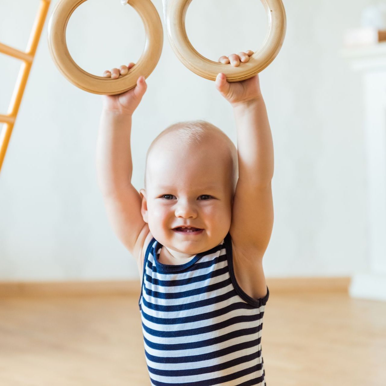 Smiling baby grasping wooden gymnastic rings, wearing a blue and white striped tank top.
