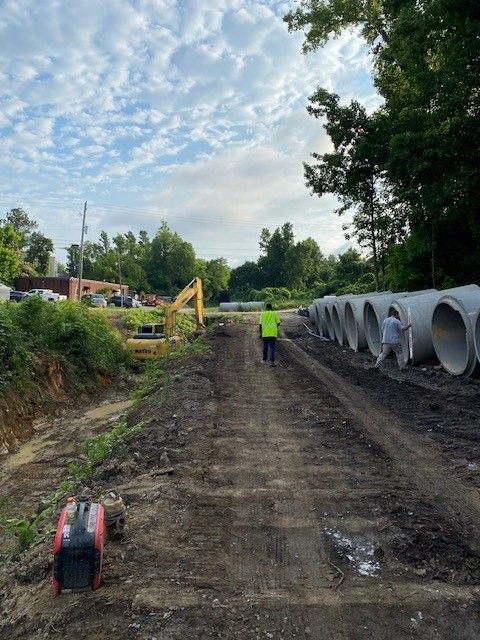 A man in a yellow vest is walking down a dirt road next to a row of pipes.