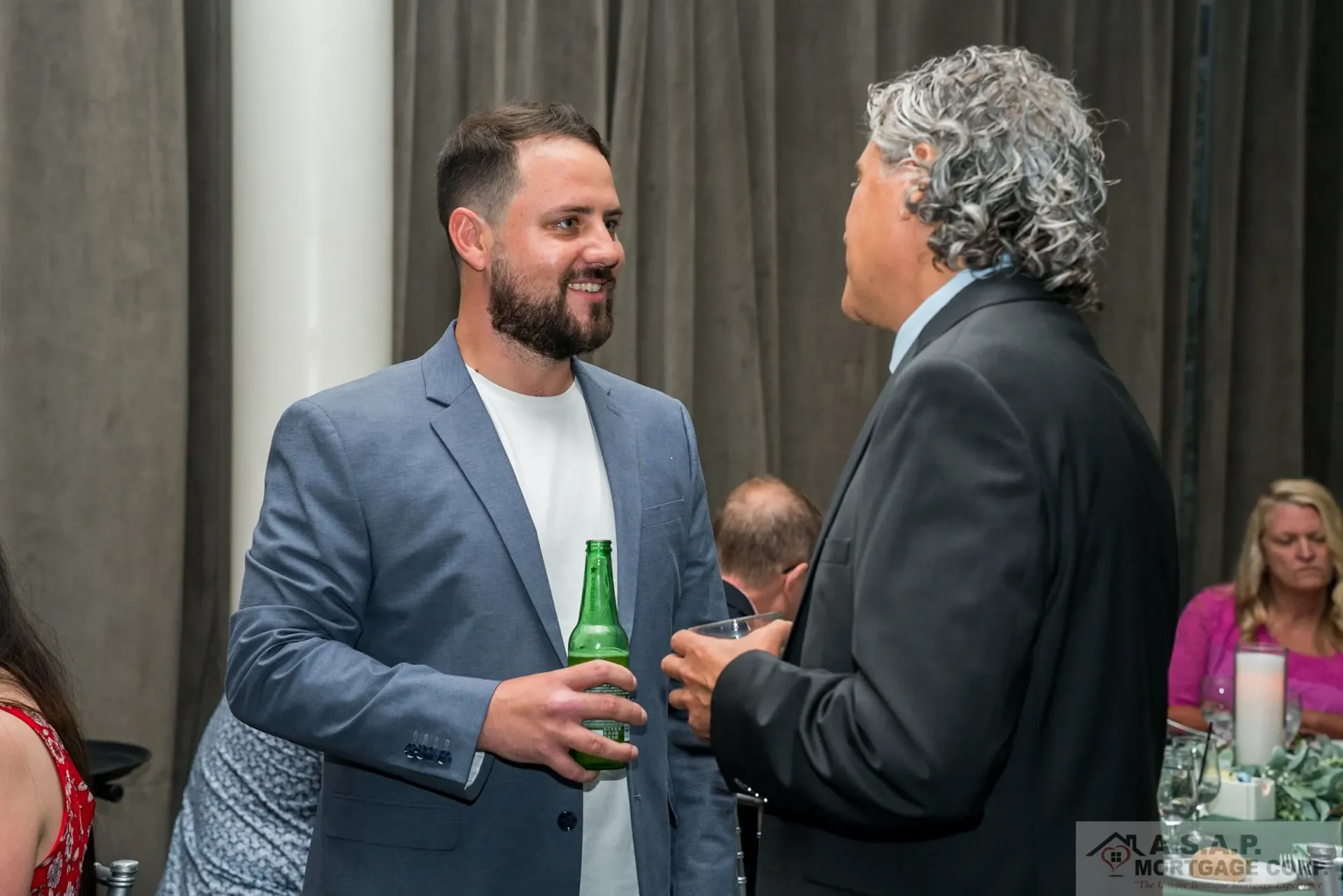 A man in a suit is talking to another man while holding a bottle of beer.