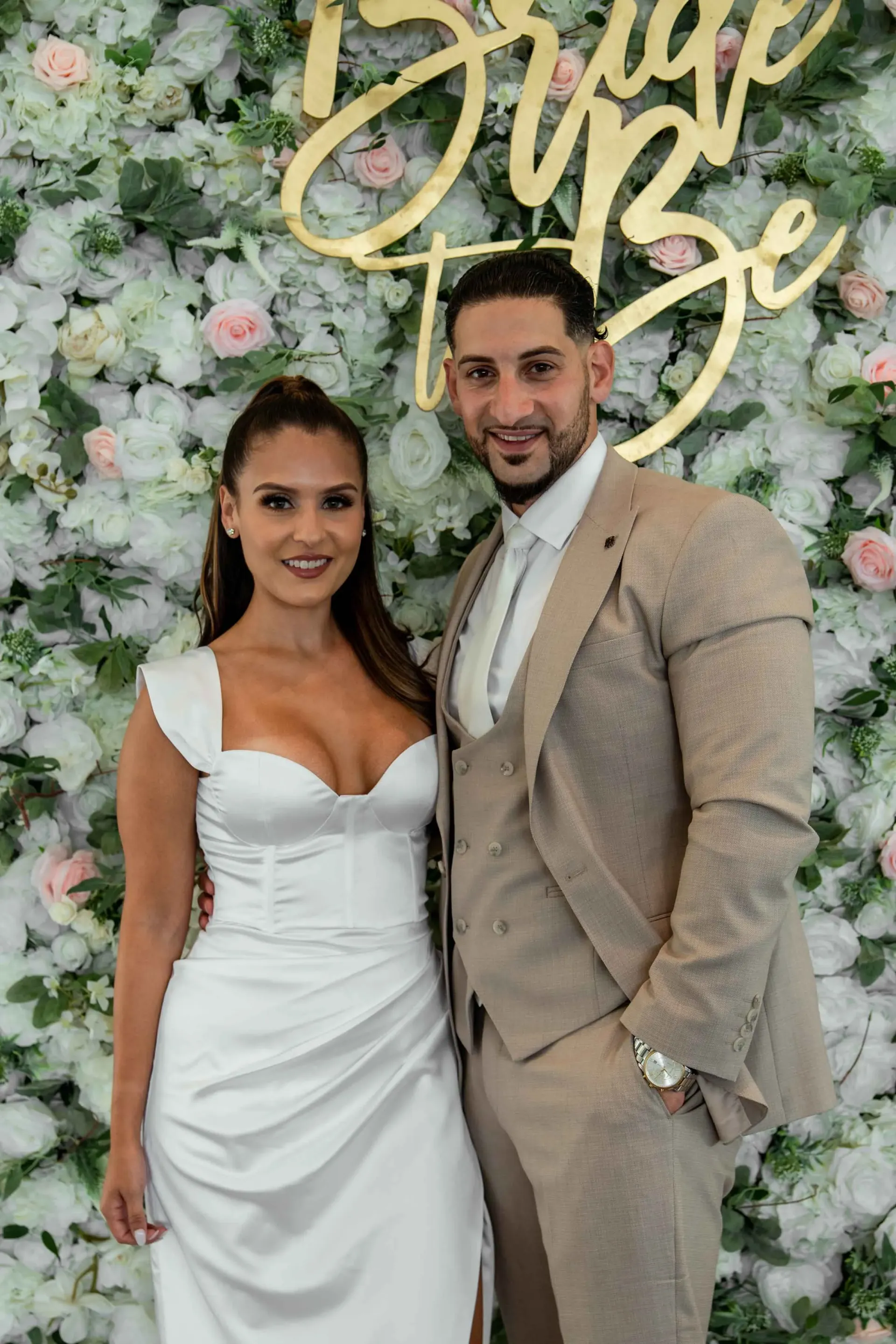 A bride and groom are posing for a picture in front of a wall of flowers.
