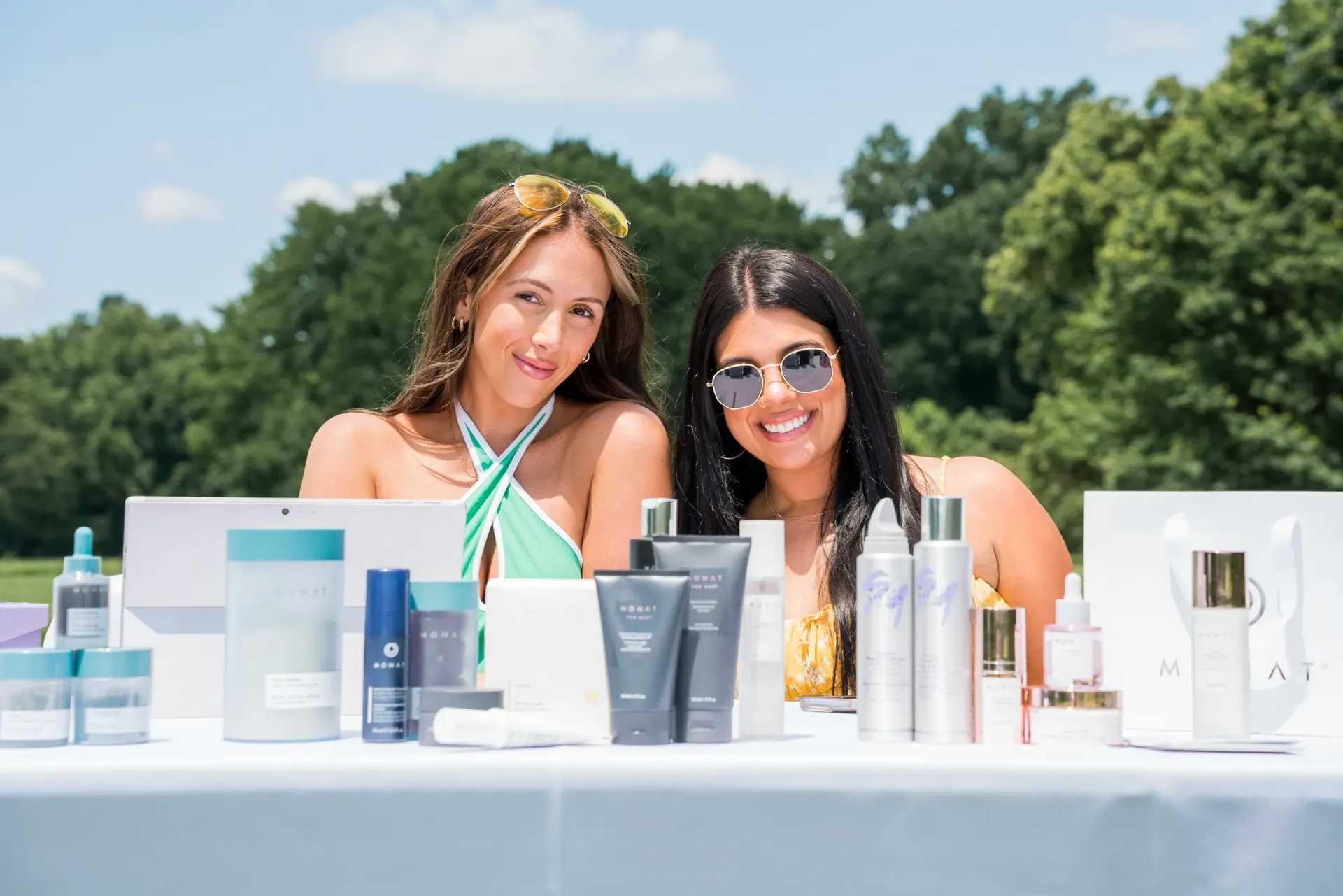 Two women are sitting at a table filled with cosmetics.