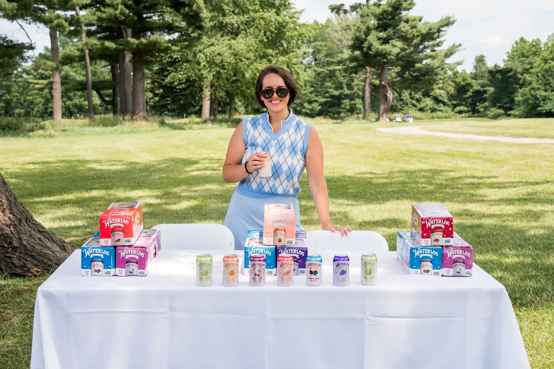 A woman is standing in front of a table filled with drinks in a park.