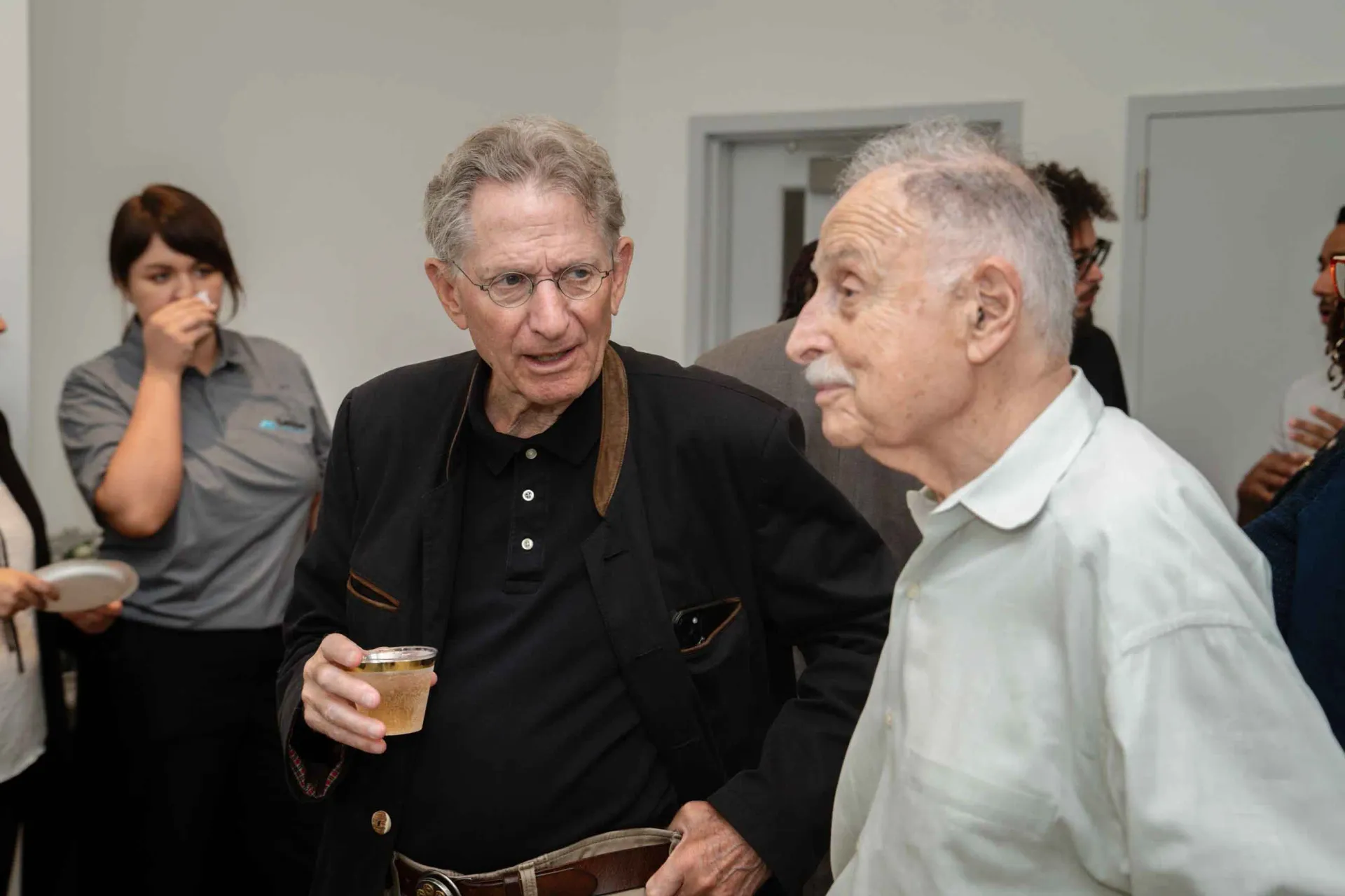 Two older men are standing next to each other in a room talking to each other.