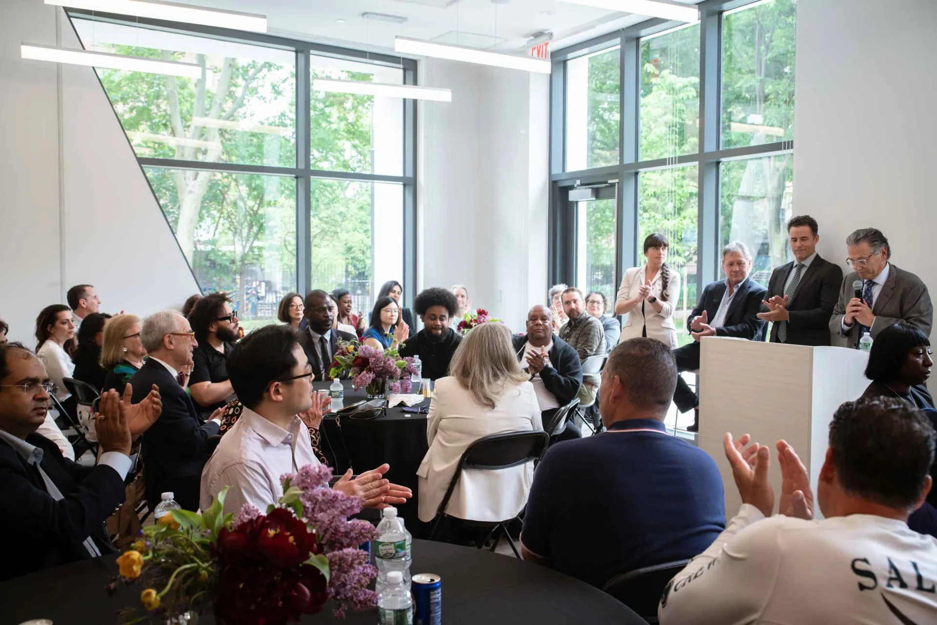 A group of people are sitting at tables in a room with a man standing at a podium giving a speech.