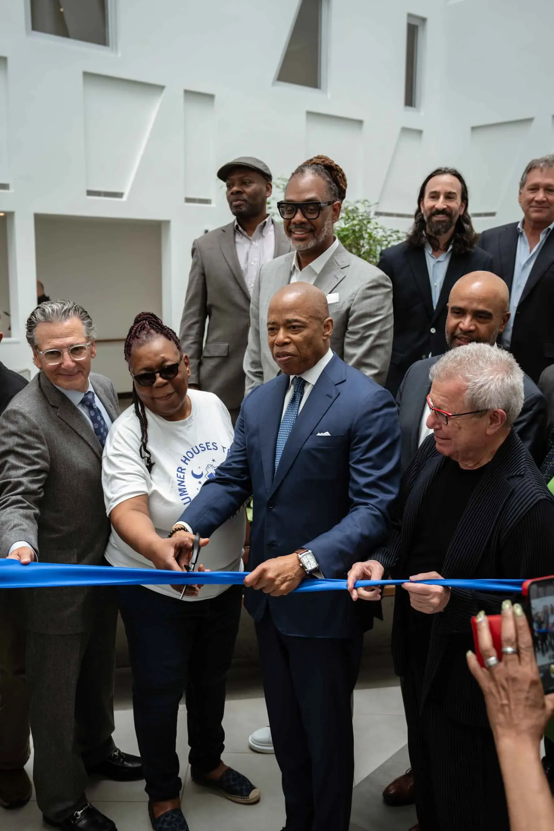 A group of people are standing around a man in a suit cutting a blue ribbon.