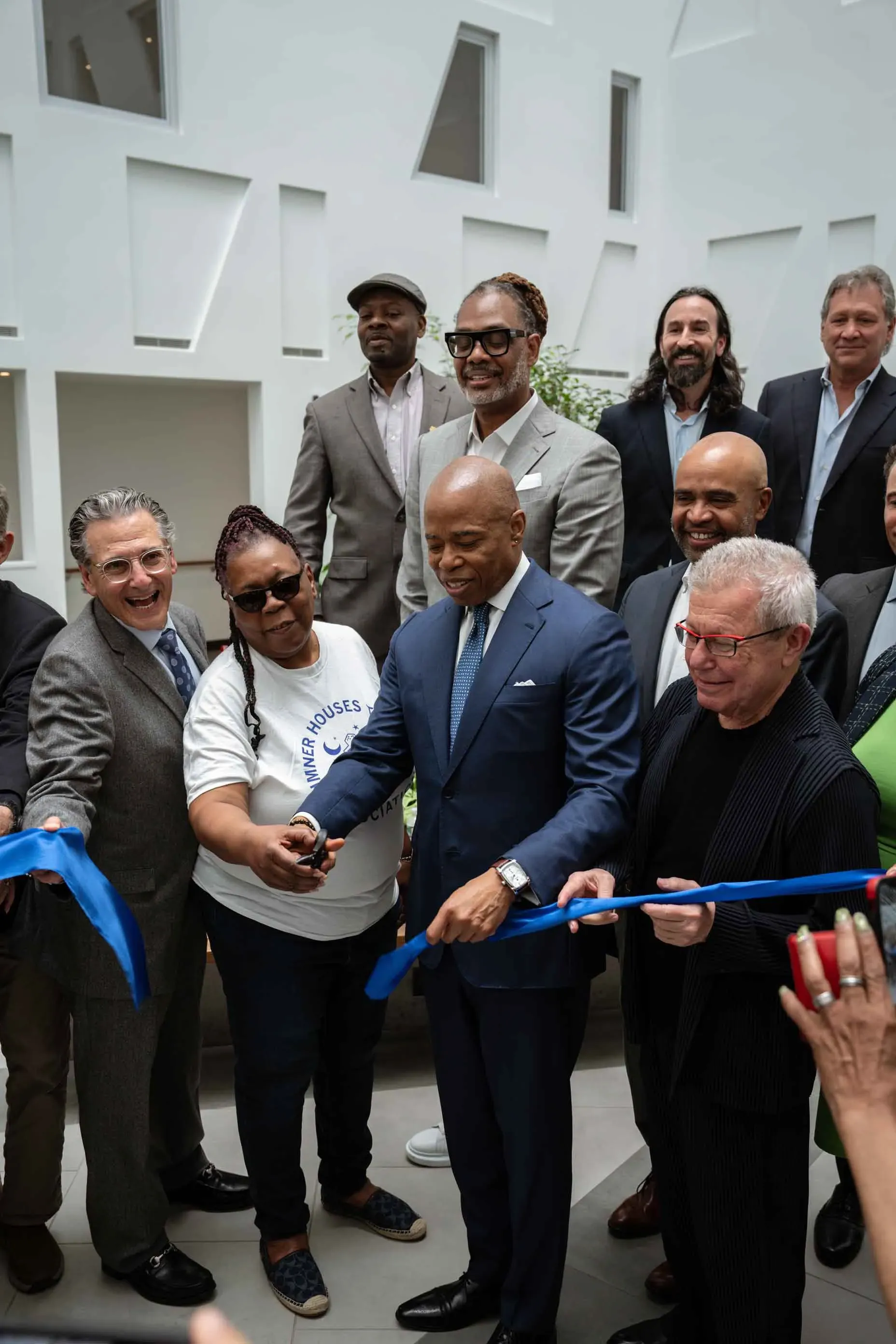 A group of people are standing around a man in a suit cutting a blue ribbon.