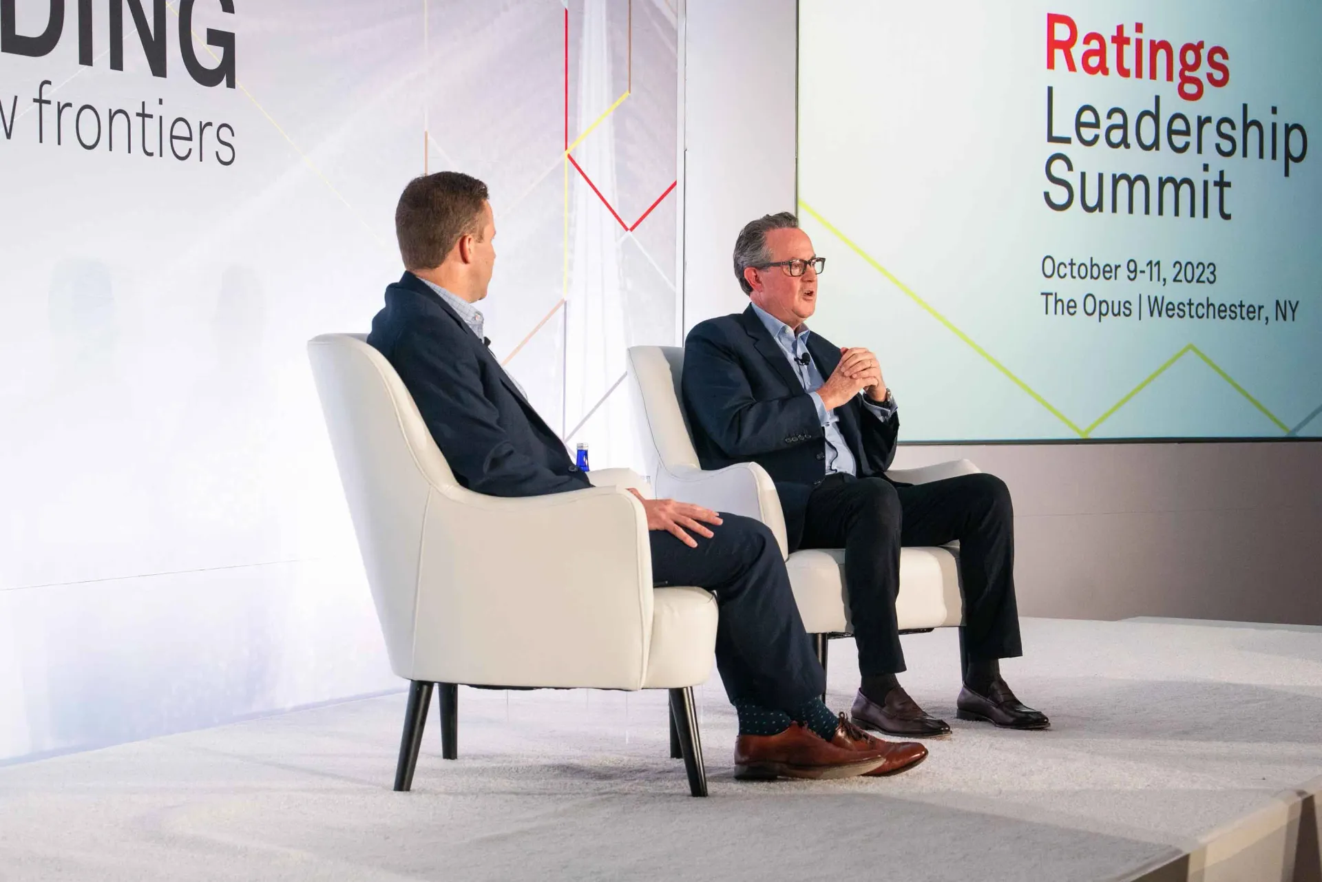 Two men are sitting in chairs on a stage at a ratings leaders summit.