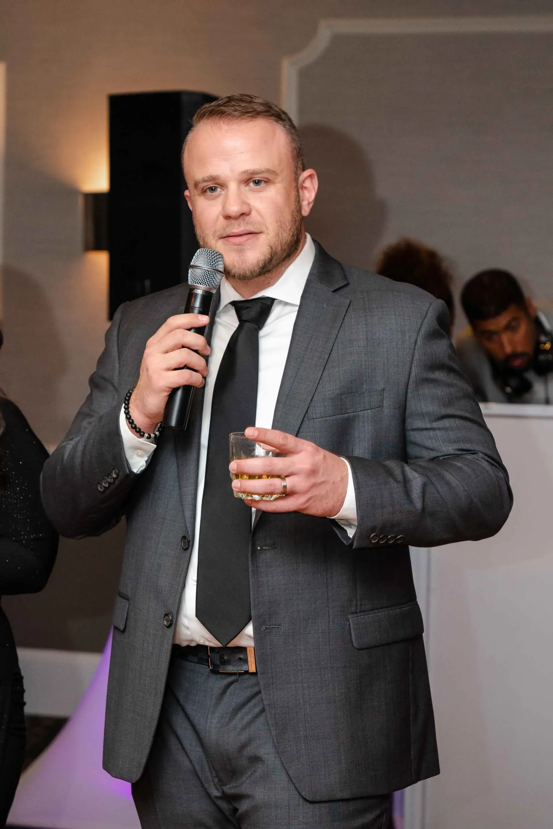 A man in a suit and tie is holding a microphone and a drink.