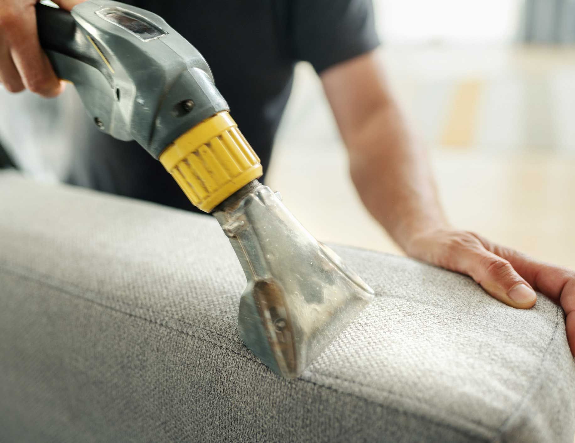A man is cleaning a couch with a vacuum cleaner.