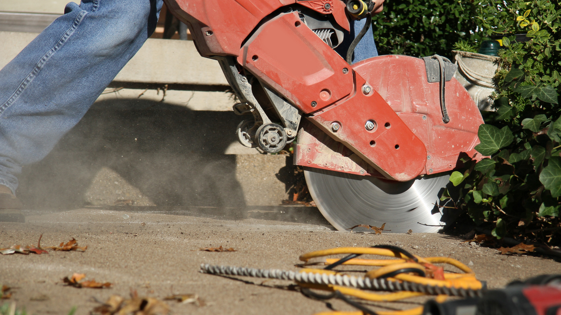 Construction worker in jeans and work boots using a concrete saw on pavement, creating a dust cloud.