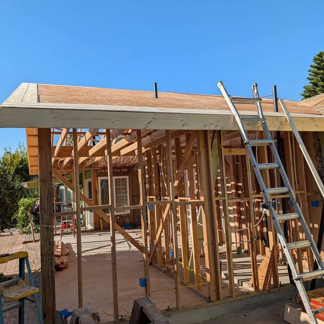 Framed house under construction with plywood roof, wooden studs, and ladders against a blue sky